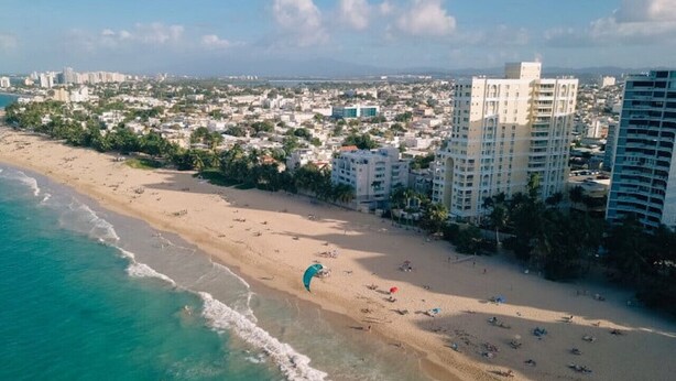 An aerial view of a beach with a parasail, buildings, and a coastline. - 2 Boho Apartments in 1 Listing: With Pool and Steps to Beach (San Juan)