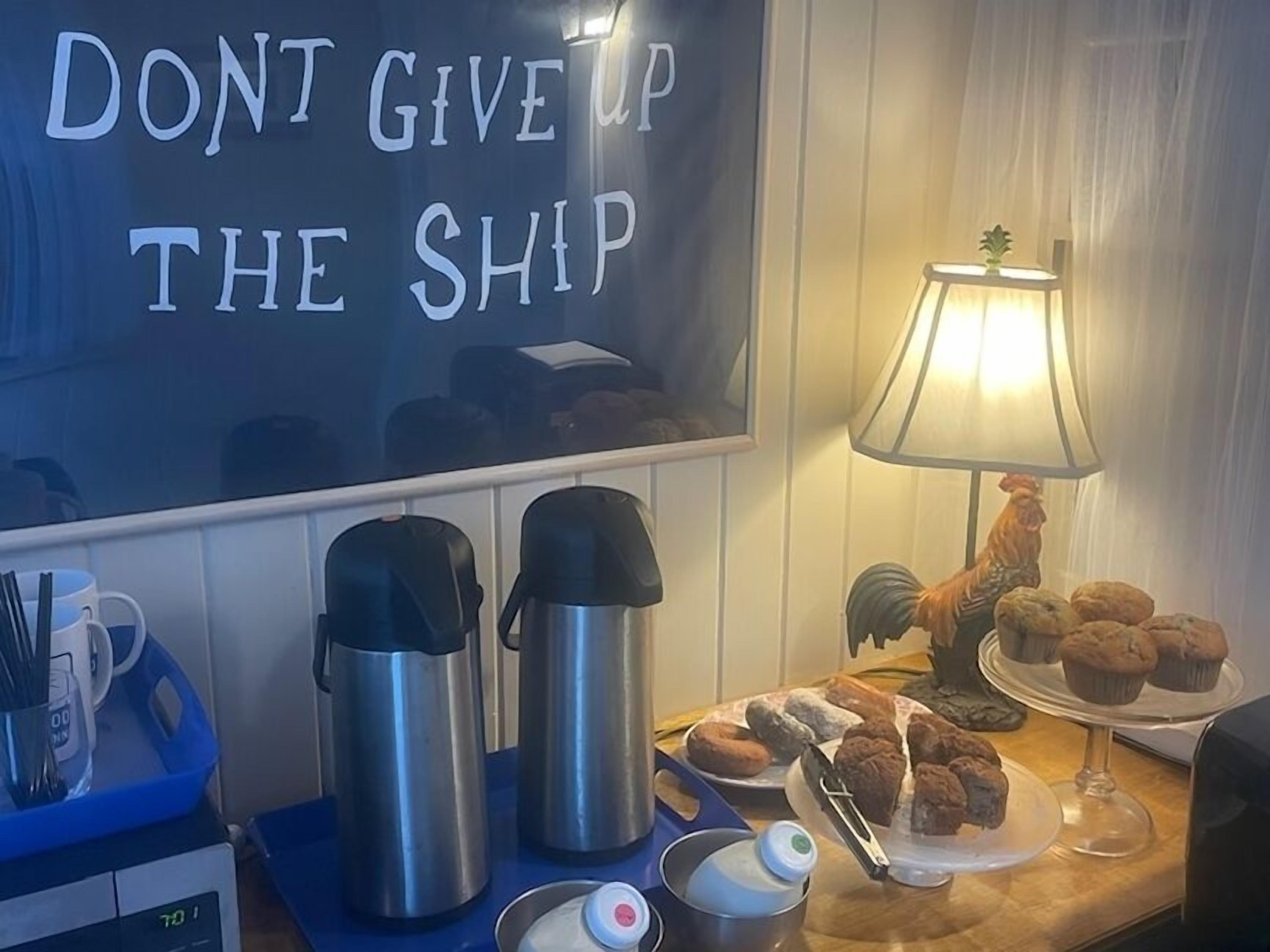 Een kitchenette met een bord waarop staat 'DON'T GIVE UP THE SHIP', roestvrijstalen waterkokers, een koffiezetapparaat, een lamp en een schaal muffins.