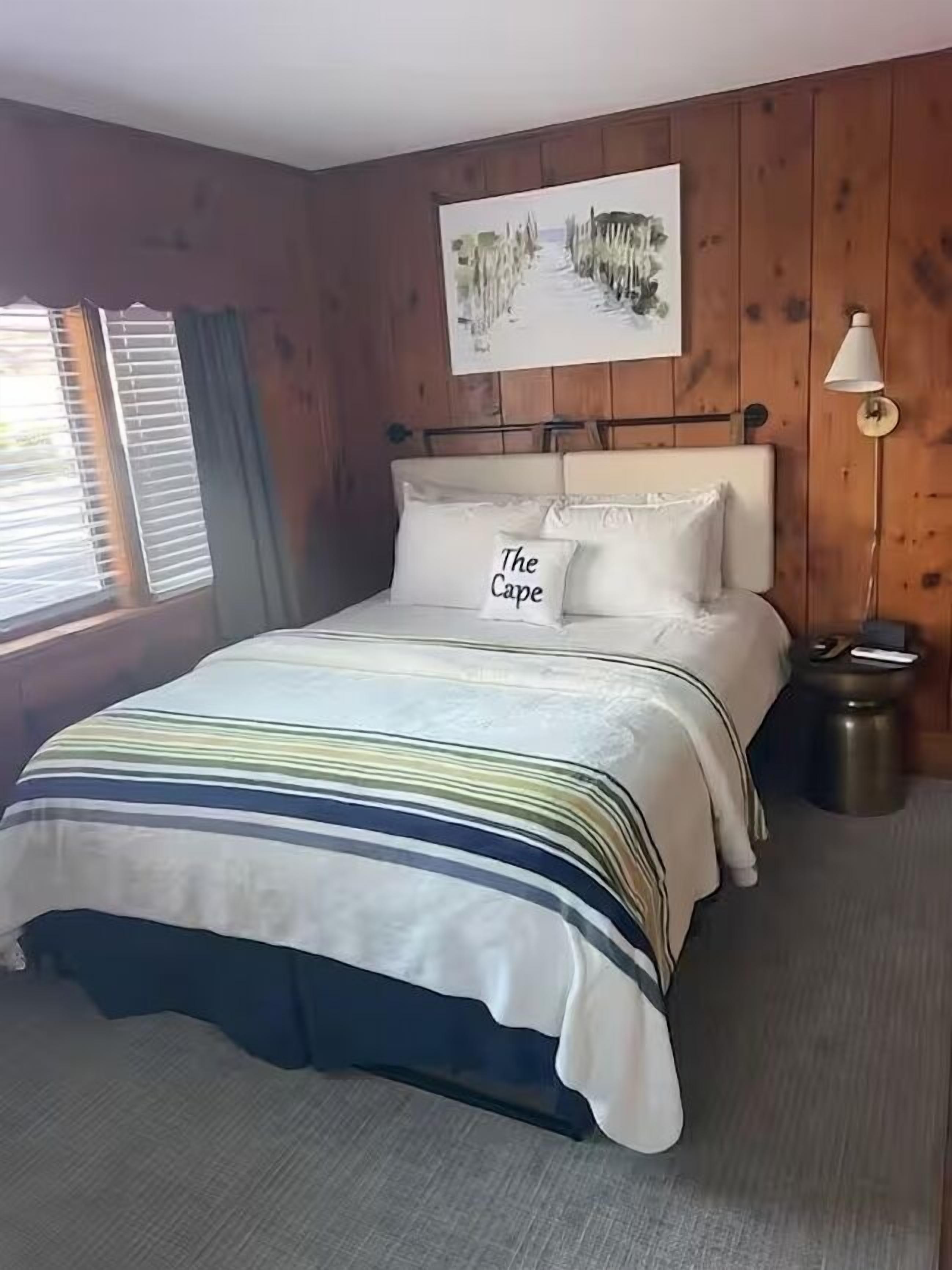 A hotel room with a bed, a painting on the wall, a bedside lamp, and a window with blinds.