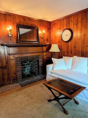 A room with wooden paneling, a fireplace, a white sofa, a lamp, and a clock on the wall. - Cape Cod Motor Inn (South Yarmouth)