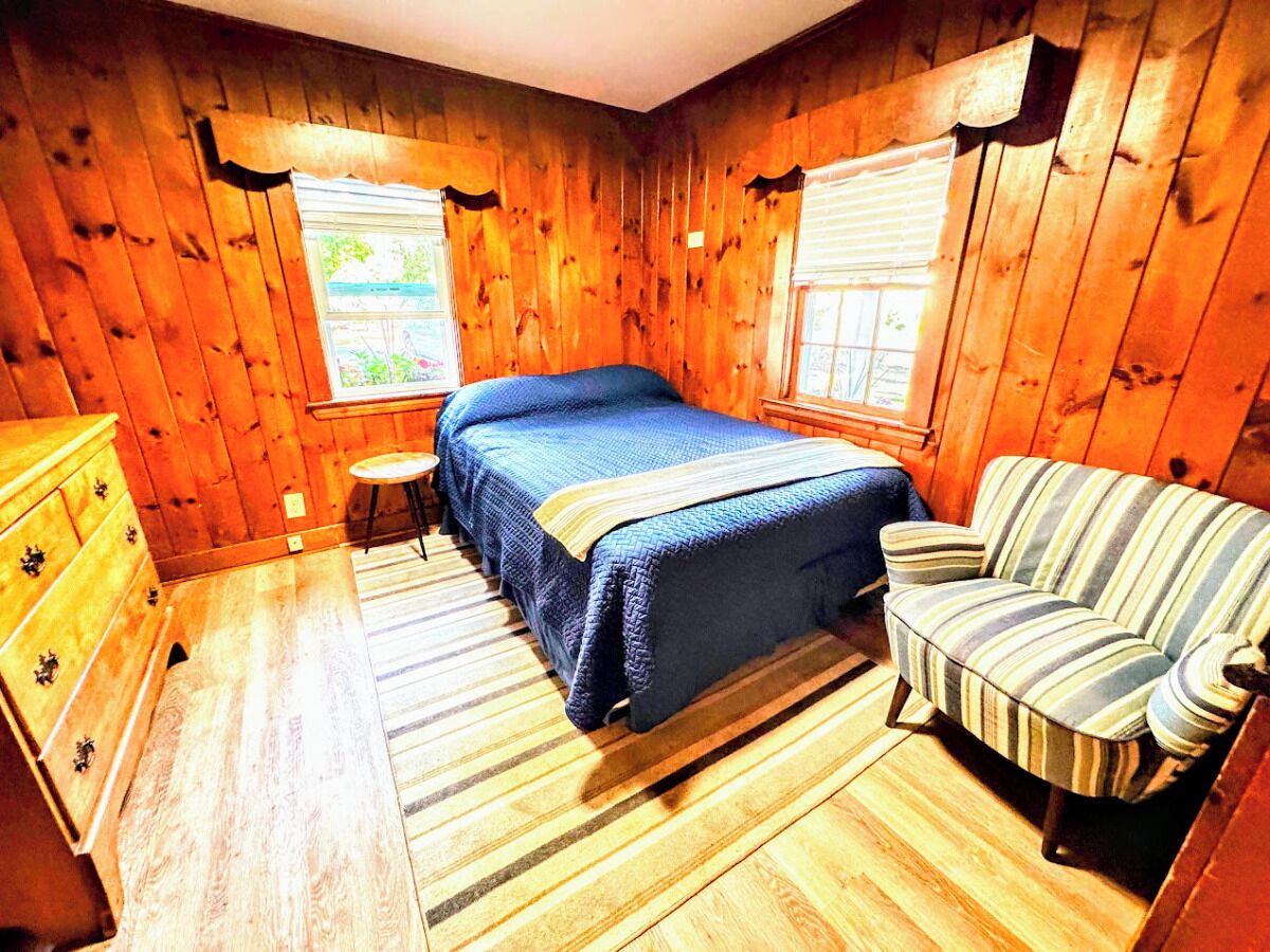 A wooden-paneled room with a bed, a striped armchair, a small table, and a dresser.