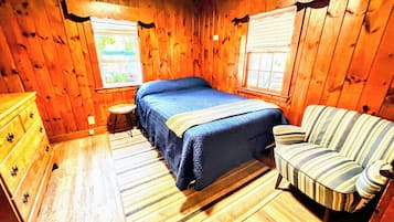 A wooden-paneled room with a bed, a striped armchair, a small table, and a dresser.