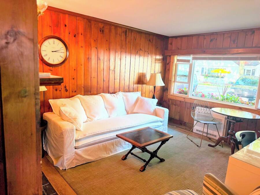 A room with a white sofa, a wooden wall, a round clock, a small table, and a window with a view of the street.