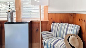 A room with a wooden wall, a striped sofa, a small refrigerator, a floor lamp, and a window with blinds. - Cape Cod Motor Inn (South Yarmouth)