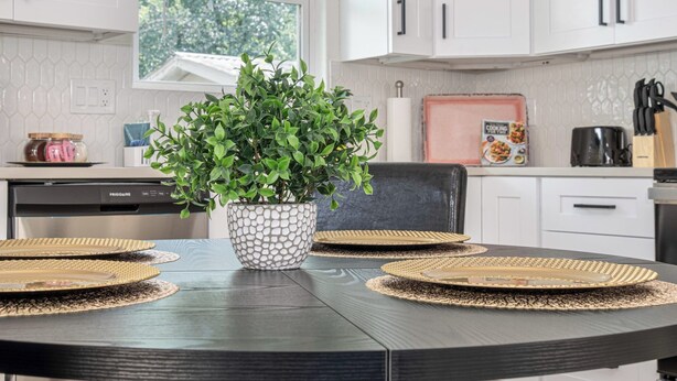 A modern kitchen with white cabinets, a black dining table with woven placemats, a potted plant, and a cooking book on the counter. - Historic Comfort 2BR/1BA Guesthouse Near Riverwalk (San Antonio)