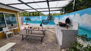 Une terrasse extérieure avec une table dressée pour les repas, un barbecue et une fresque représentant une scène de plage.