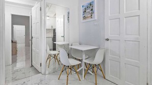 A modern dining area with white tables and chairs, a kitchen in the background, and a door leading to another room. - Coastal Pleasures Studio (Saint Ann's Bay)