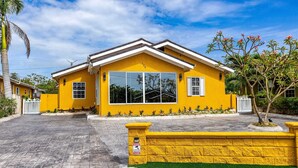 A yellow two-story house with a large window, a paved driveway, and a small tree in the front yard. - Coastal Pleasures Studio (Saint Ann's Bay)