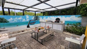 Un patio exterior con mesa, sillas, una parrilla y un mural de una playa.