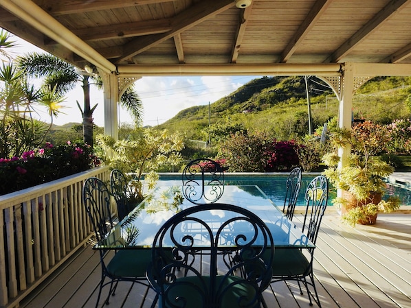 A covered outdoor area with a glass table and chairs, surrounded by lush greenery and a pool.