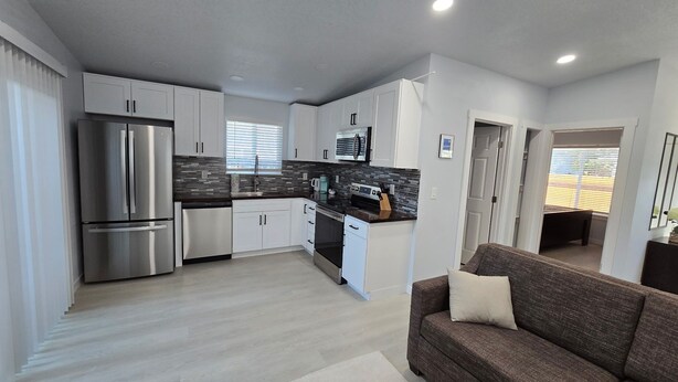 A modern kitchen with white cabinets, a stainless steel refrigerator, and a microwave. There is a brown sofa and a small rug in the living area. - One bedroom house (Portland)