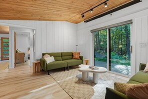 A modern living room with a green sofa, a white coffee table, and a large sliding glass door leading to a forested area. - Luxury Scandinavian nature escape, private saun (Springfield)