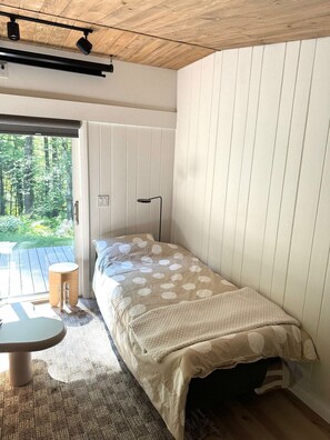Una cama individual con colcha estampada, una mesita redonda pequeña, una lámpara de pie y vistas a un bosque a través de una puerta corrediza.