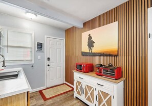 A compact kitchen with a microwave and toaster on a wooden cabinet, a door with a welcome sign, and a wall-mounted painting of a sunset. - Vibrant Cowgirl Hideaway - Rodeo Themed, Fire Pit & 1/2 mile walk to Whiskey Row (Prescott)