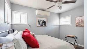 Ein Schlafzimmer mit einem Bett, einem Deckenventilator, zwei Fenstern mit Jalousien und einem kleinen Tisch mit Kissen.