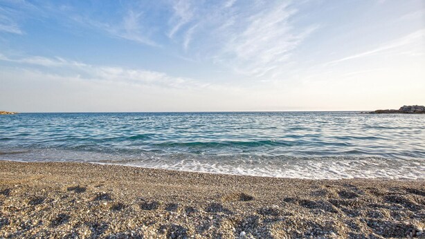 Una platja de petits trossos de pedra amb aigua blau clar i un promontori rocosós a la distància.