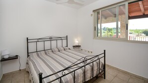Ein Schlafzimmer mit Bett, Nachttisch, Lampe, Deckenventilator und Fenster mit Blick auf Grünflächen.
