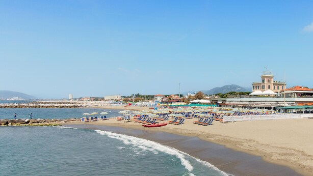 Una playa con filas de tumbonas y sombrillas, un muelle y un edificio con una torre.