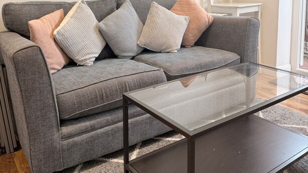 A grey sofa with multiple cushions, a glass coffee table, and a large mirror on the wall. - Cosy Adorable 1 Bedroom Studio in London (London)