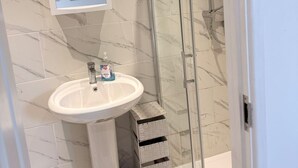 A bathroom with a marble-tiled wall, a white pedestal sink, a glass shower enclosure, and a framed picture above the sink. - Cosy Adorable 1 Bedroom Studio in London (London)