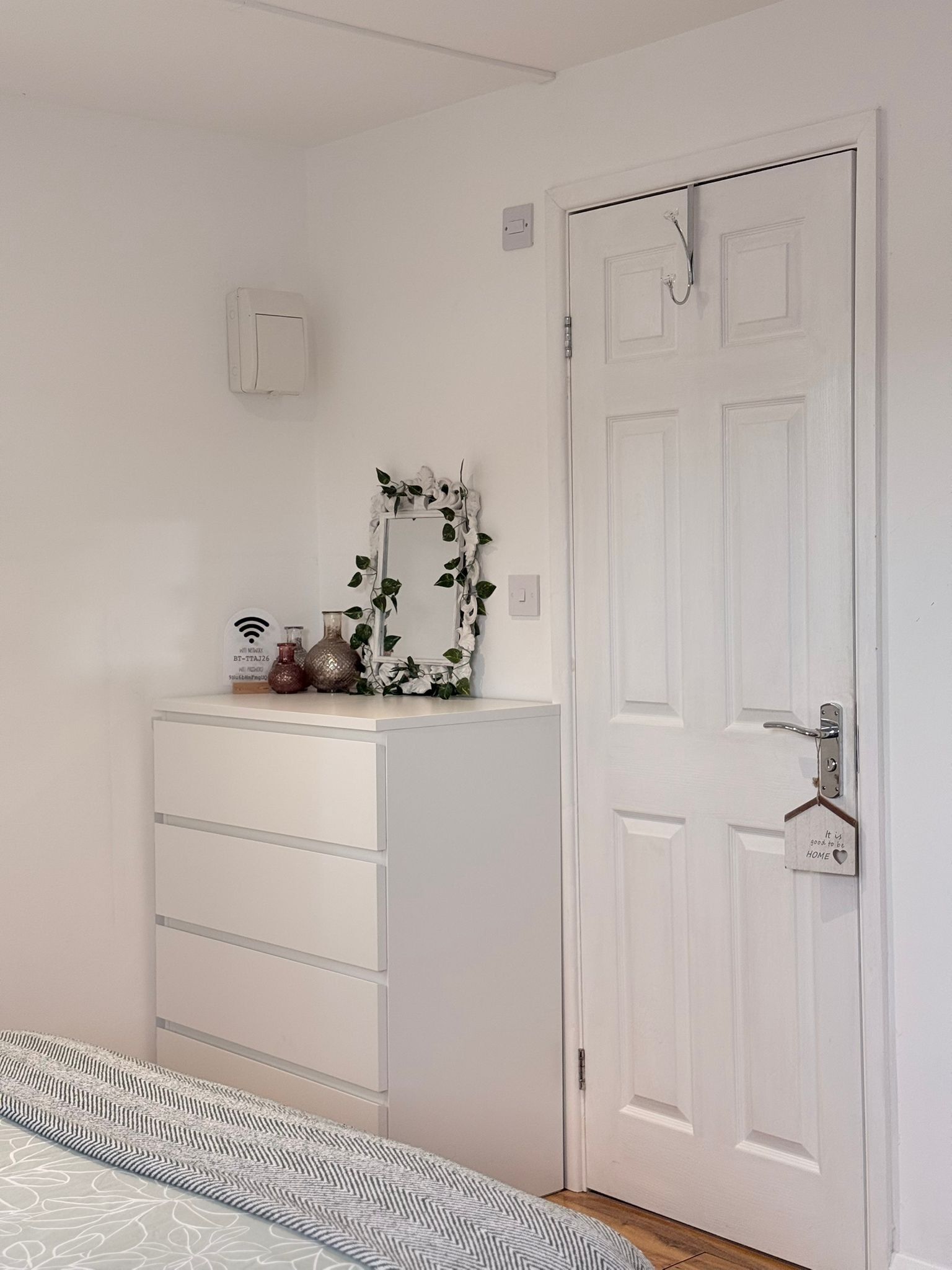 A bedroom with a white dresser, a mirror with a leaf frame, a door with a heart-shaped lock, and a patterned bedspread.