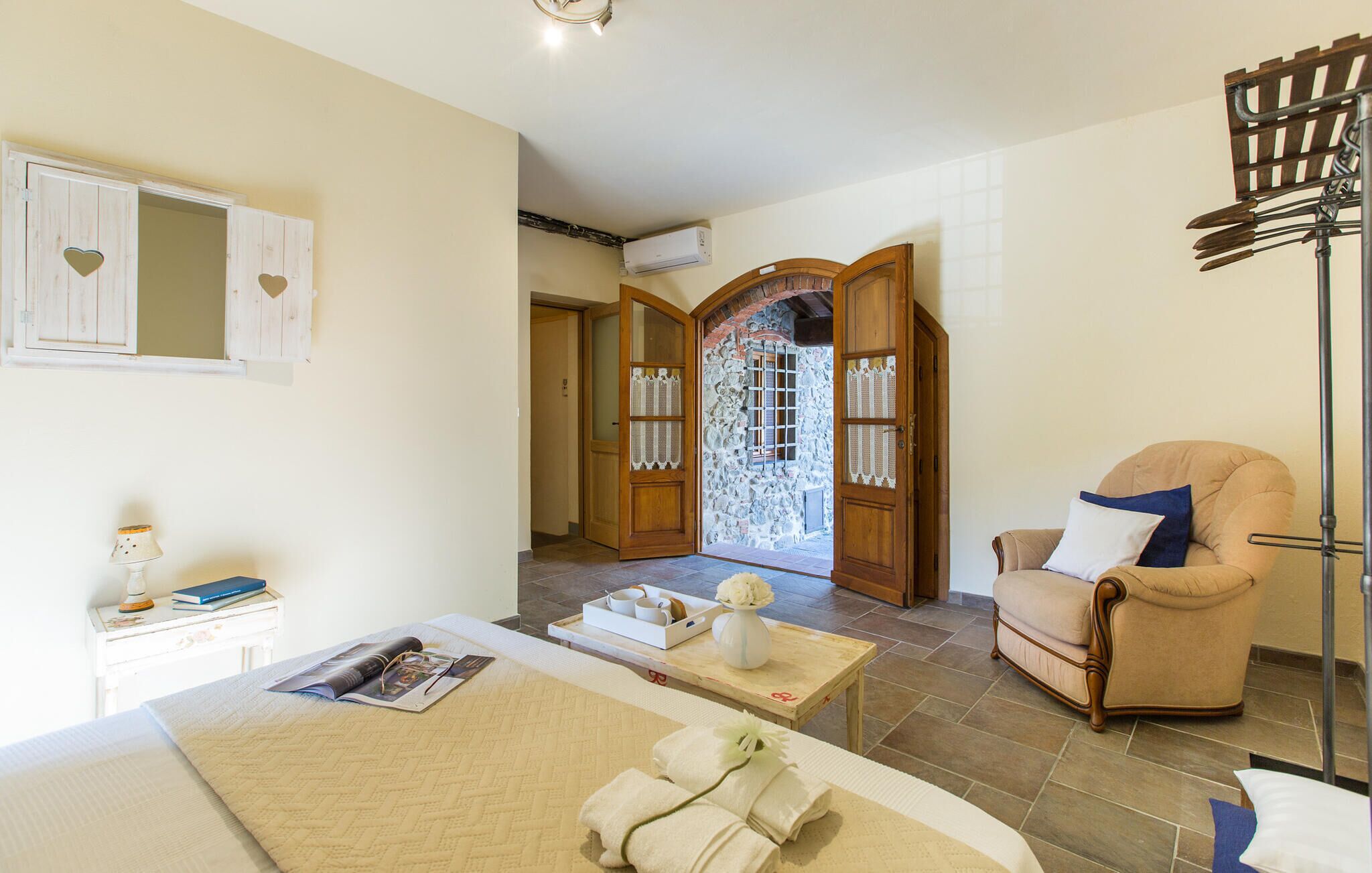Habitación de hotel con cama, una silla, una mesa con bandeja de toallas y pared de piedra con puerta de madera.