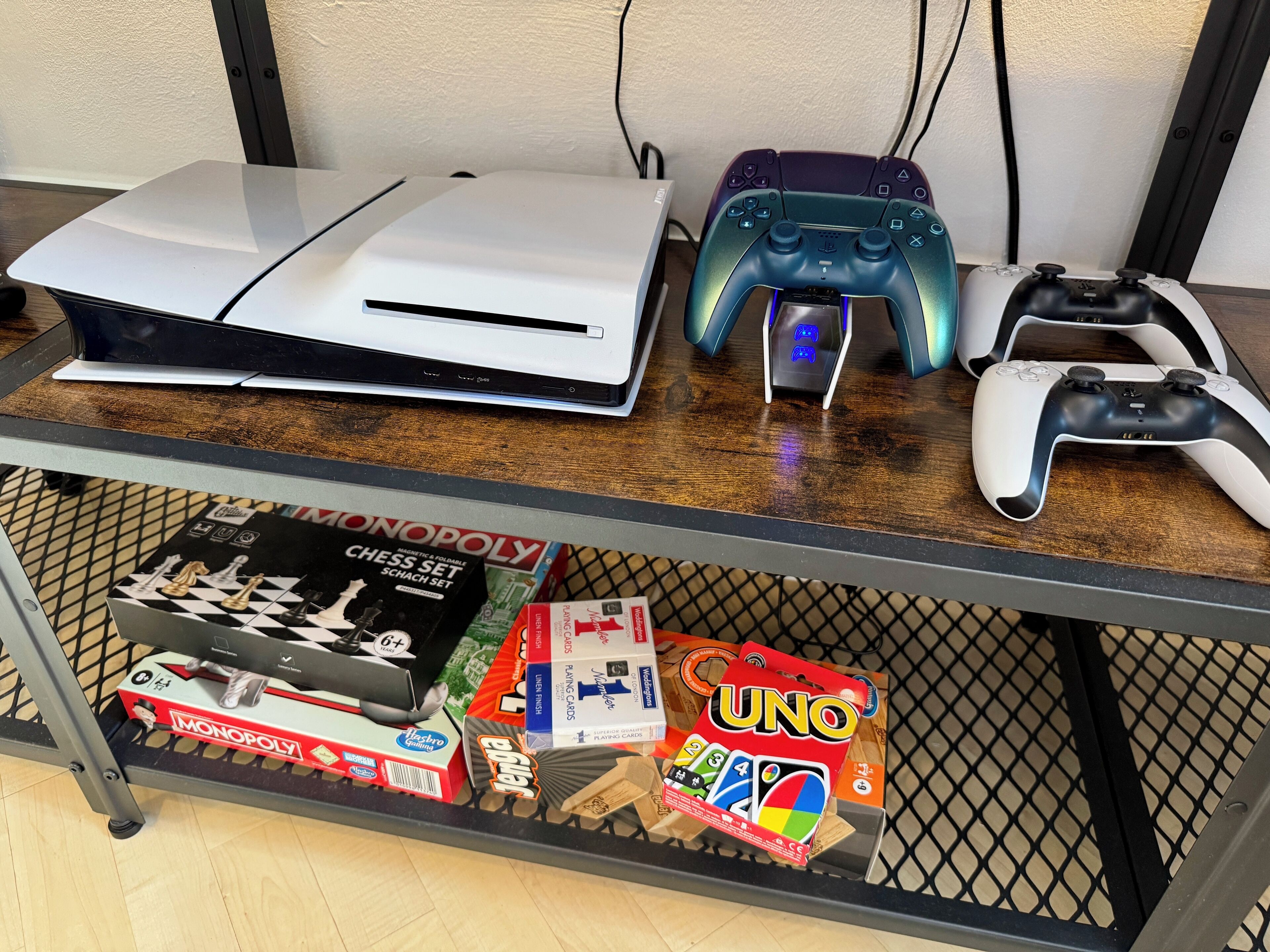 Peliasennus, jossa on PlayStation 5 -pelikonsoli, kaksi peliohjainta ja lautapelejä, kuten Monopoly ja UNO, hyllyssä.