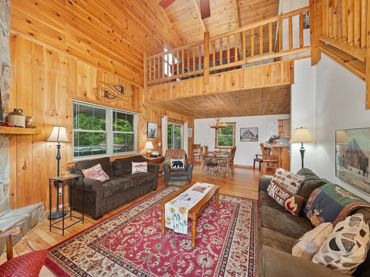 A spacious living room with a high ceiling, wooden walls, and a balcony. There are two sofas, a coffee table, and a dining area with chairs.