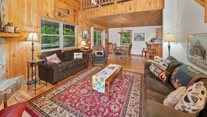 Ein geräumiges Wohnzimmer mit hohen Decken, Holzwänden und einem Balkon. Es gibt zwei Sofas, einen Couchtisch und einen Essbereich mit Stühlen.