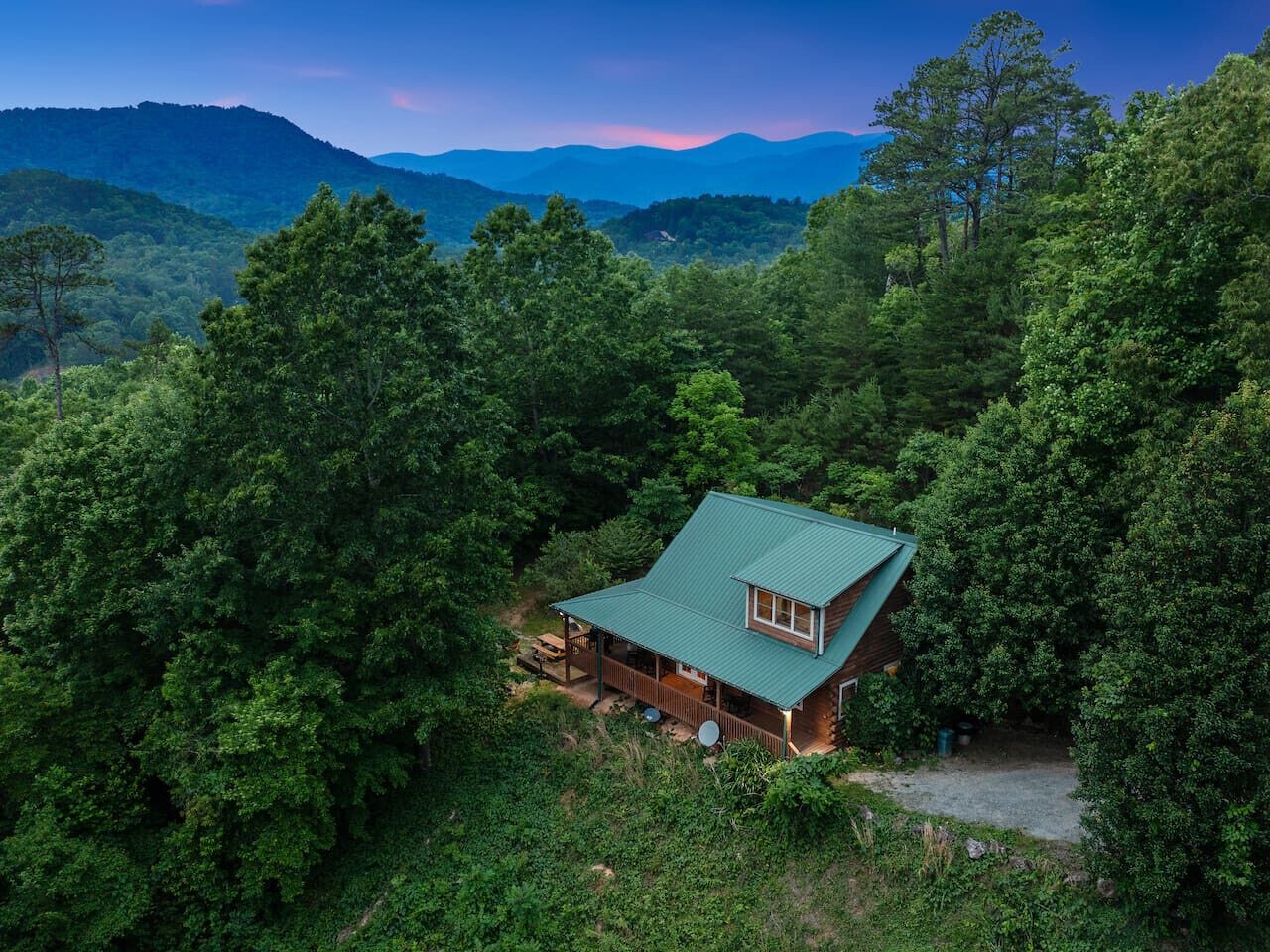 A cabin with a green roof surrounded by dense forest, overlooking a range of mountains.