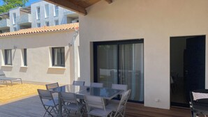 Eine überdachte Terrasse im Freien mit einem Glastisch und Stühlen, ein Holzdeck und Blick auf angrenzende Gebäude.