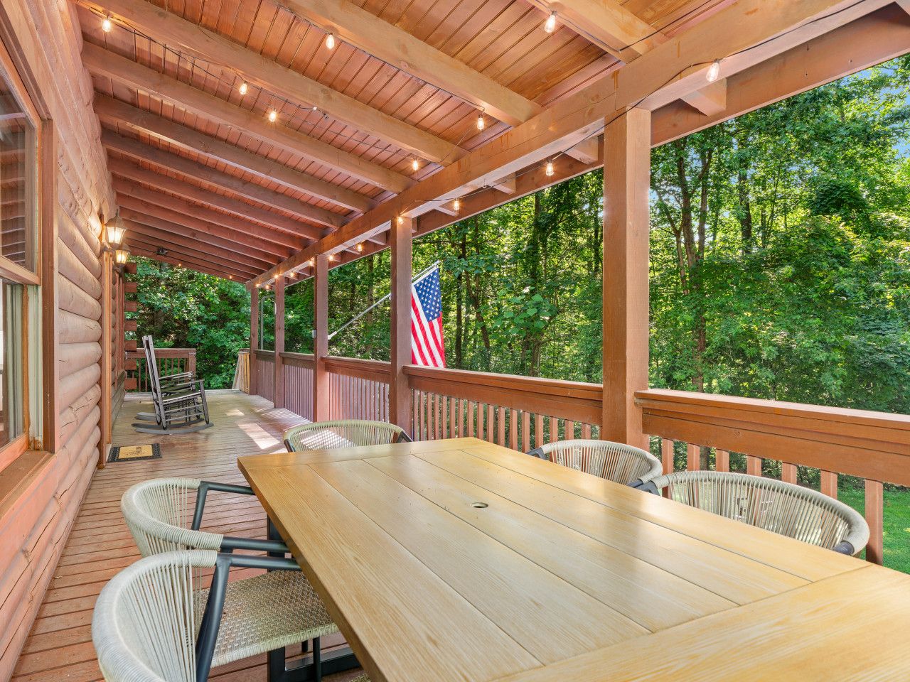 Une terrasse extérieure couverte, avec une longue table et des chaises en bois, un drapeau américain et une vue sur une zone boisée.