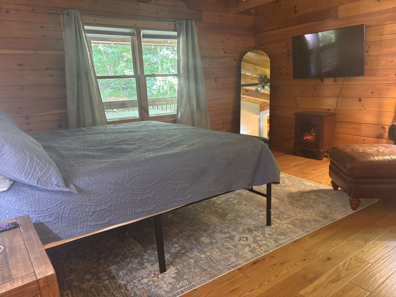 A wooden cabin bedroom with a bed, a television, a fireplace, and a large window with curtains.
