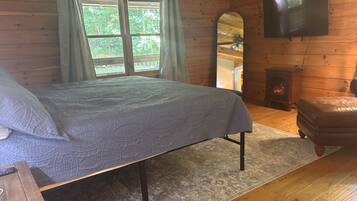 A wooden cabin bedroom with a bed, a television, a fireplace, and a large window with curtains.