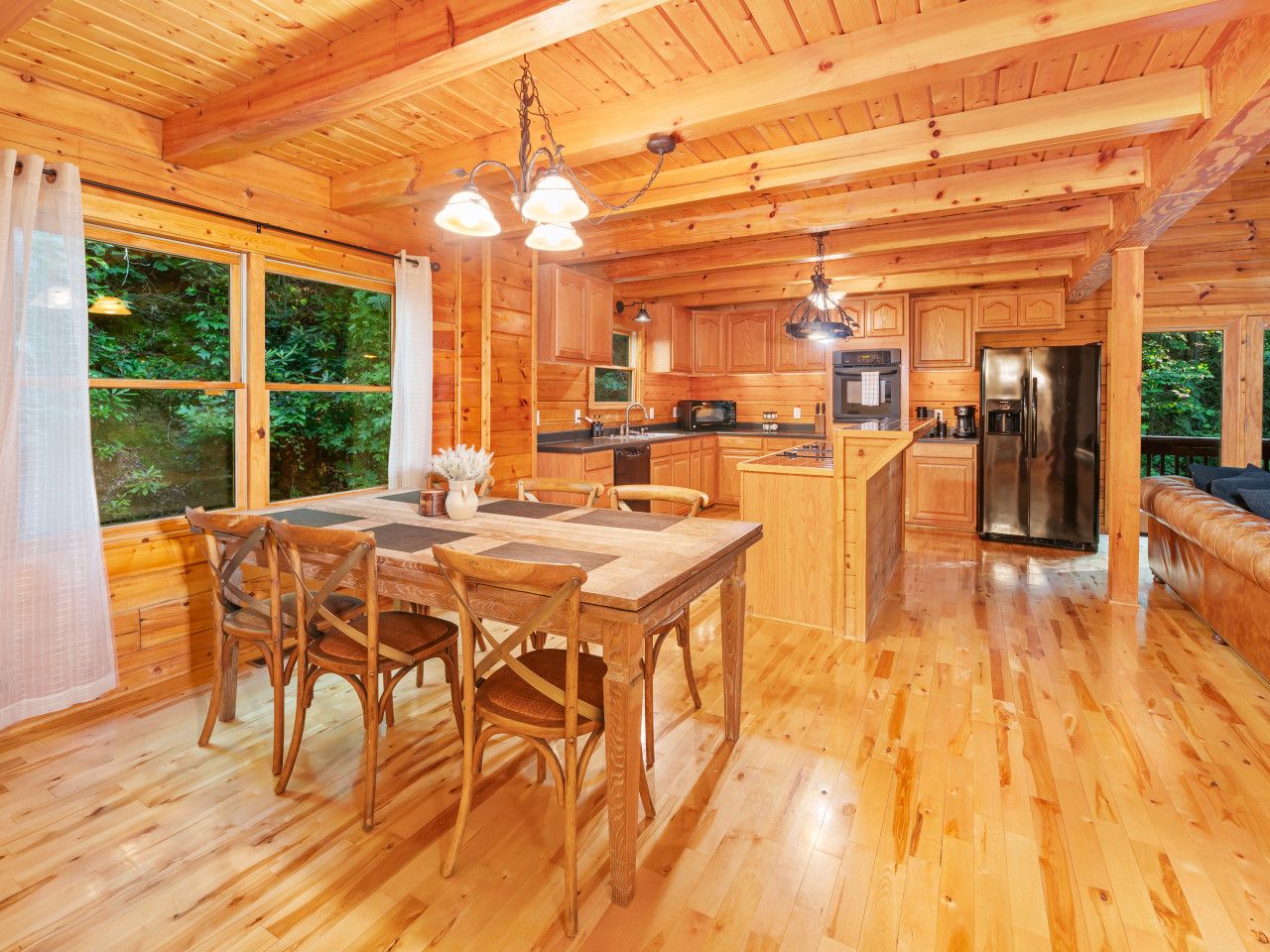 A wooden cabin interior with a dining area, kitchen, and a large window overlooking greenery.