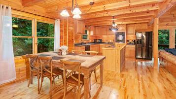 A wooden cabin interior with a dining area, kitchen, and a large window overlooking greenery.