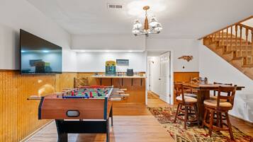 A cozy living room with a foosball table, a dining area with wooden chairs, a kitchen area with a bar, and a staircase.