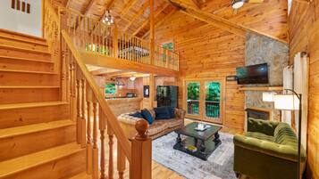 A wooden cabin interior with a staircase, a living room with a sofa and coffee table, a fireplace, and a television.