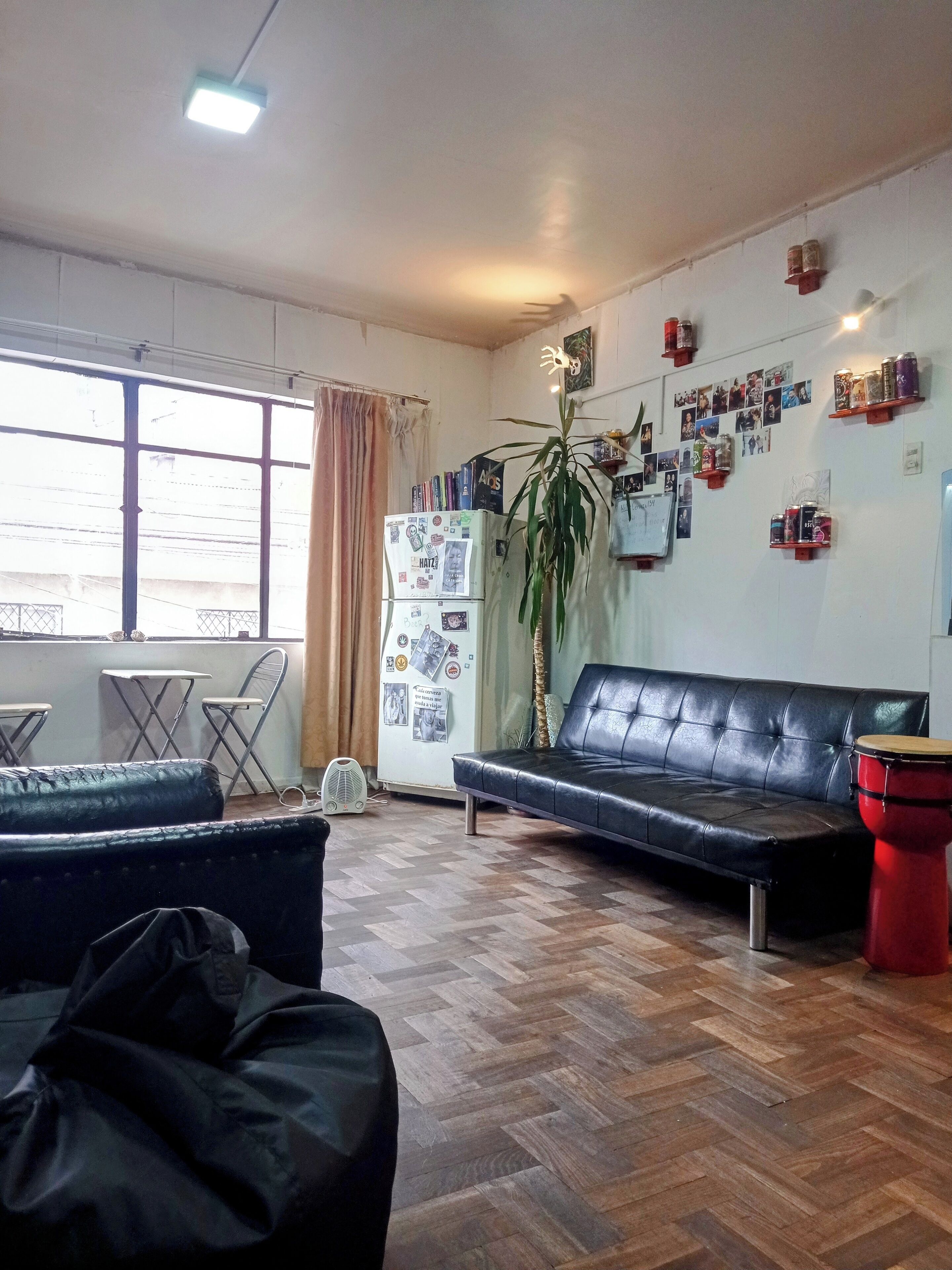 A living room with a sofa, a red drum, a refrigerator with photos, and a large window.