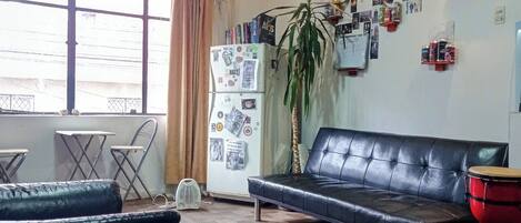 Una sala de estar con sofá, un tambor rojo, un refrigerador con fotos y un ventanal grande.