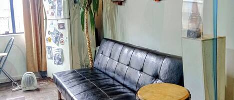 Quarto com sofá de couro preto, um tambor vermelho, geladeira, ventilador e parede decorada com fotos e latas.