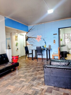 Un salón con pared azul, suelo de madera, sofá de cuero negro, un tambor rojo y una mesa de comedor con sillas.