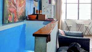 A room with a blue wall, a painting, a television, a refrigerator, a desk, and a window with curtains. - Beer and Beer (Valparaíso)