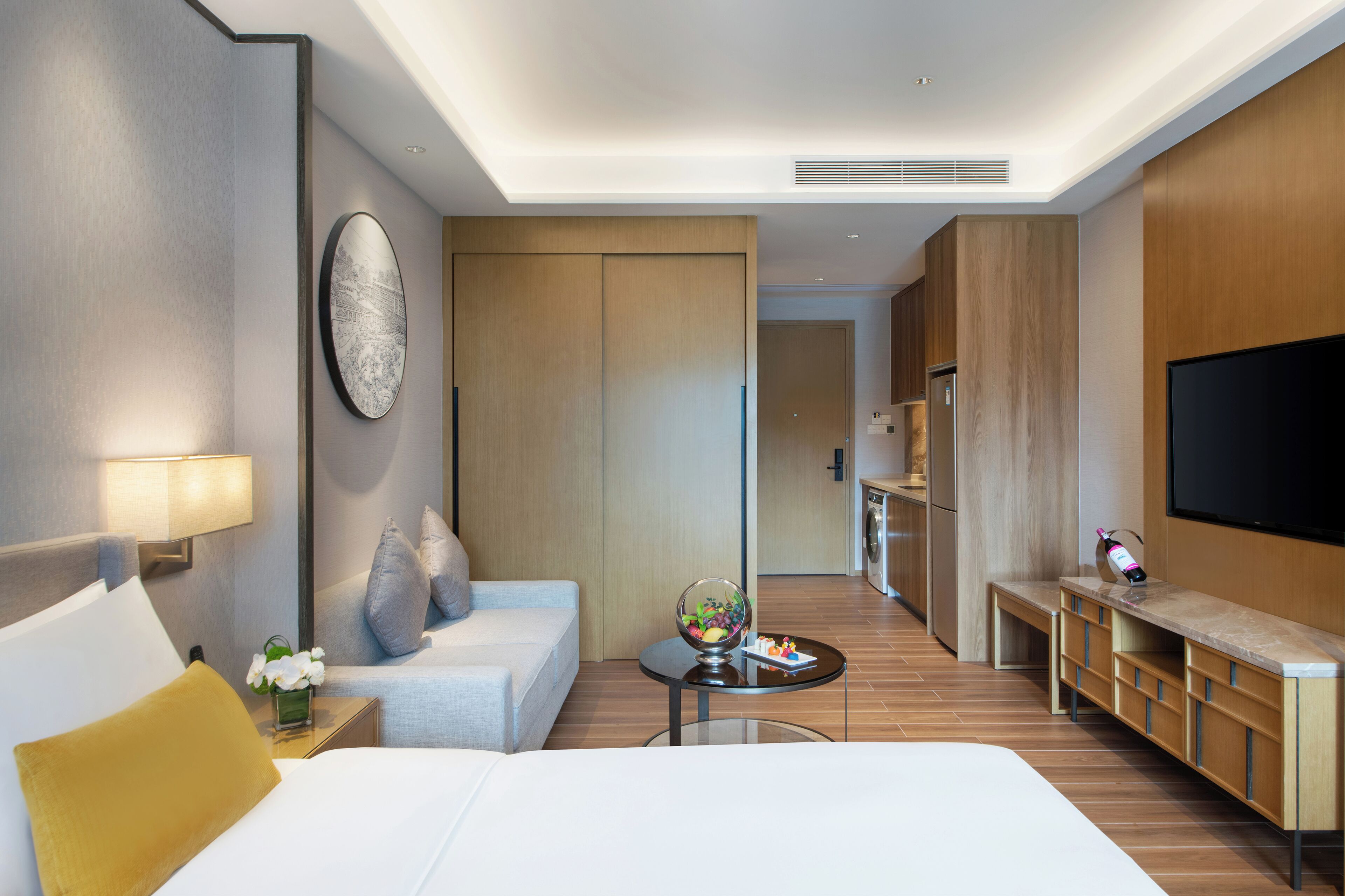 A modern hotel room with a bed, a small table with a globe, a sofa, a TV, and a kitchenette area.
