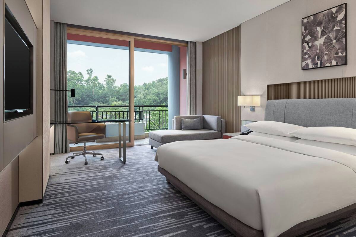 Ein modernes Hotelzimmer mit einem großen Bett, einem Schreibtisch mit Stuhl, einem Balkon mit Blick auf Bäume und einem Wandbild.