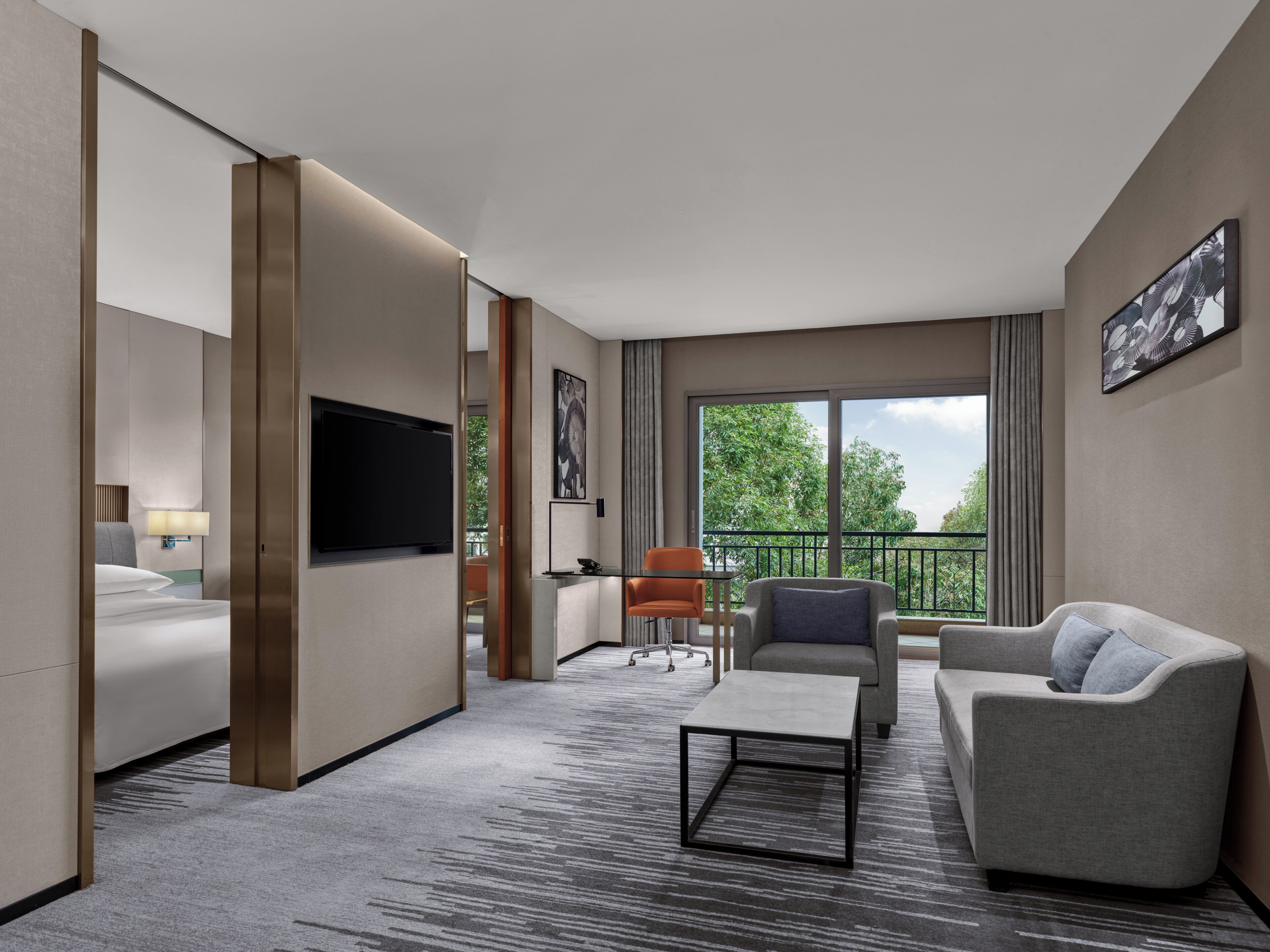 A modern hotel room with a bed, a TV, a seating area with a sofa and armchair, and a balcony with a view of trees.