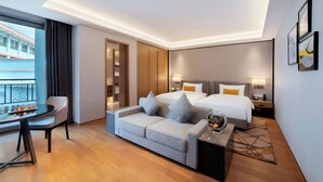 Una habitación de hotel moderna con sofá, una mesa de comedor, una cama con dos almohadas, una mesita de noche y una ventana con vistas a un edificio.
