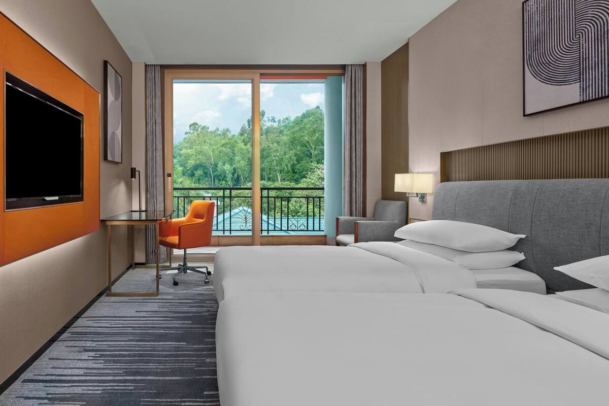 Ein Hotelzimmer mit einem großen Bett, einem Schreibtisch mit Fernseher, einem orangefarbenen Sessel, einem grauen Ohrensessel und einem Balkon mit Blick auf Bäume.