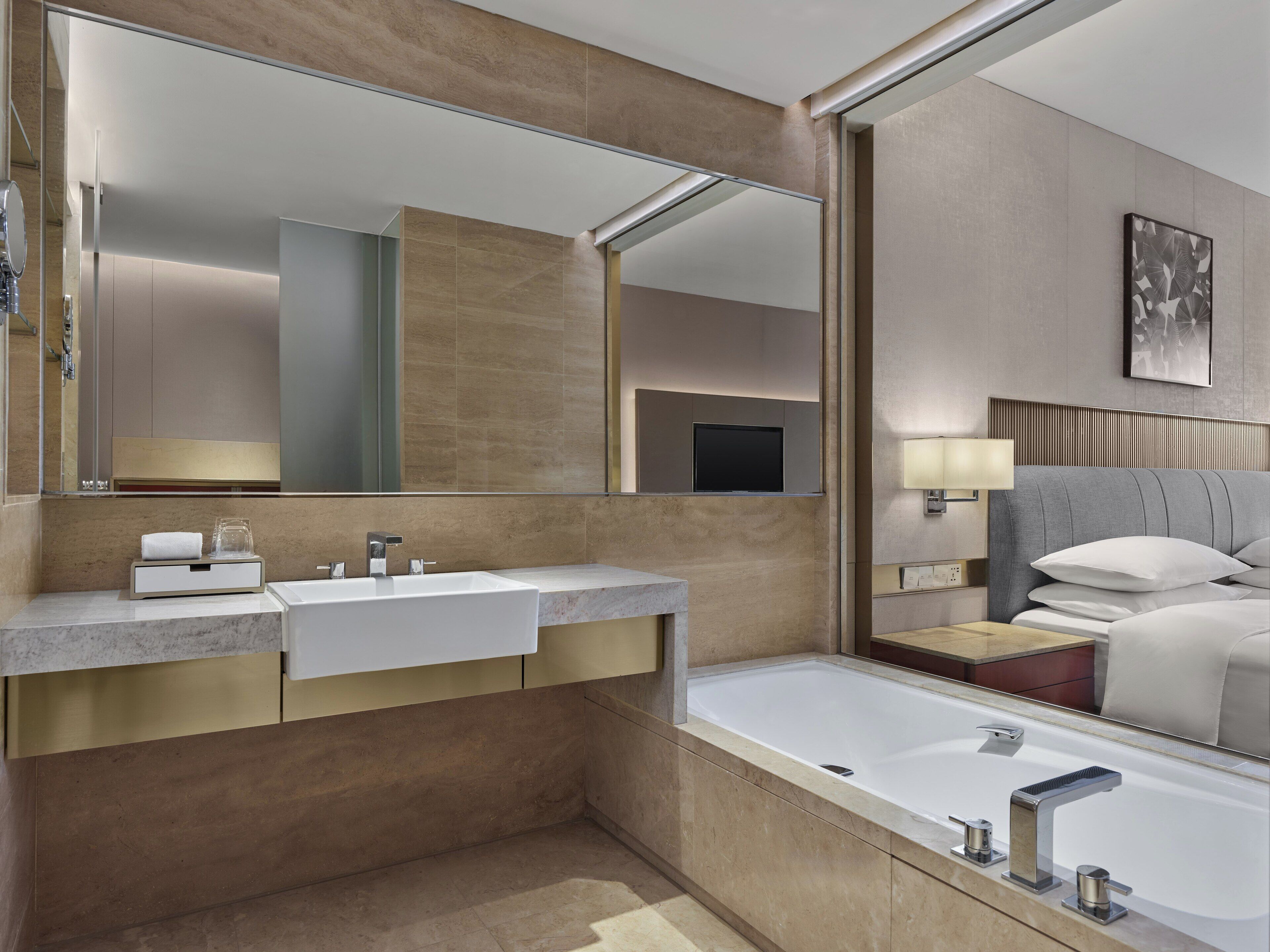 A modern bathroom with a large mirror, two sinks, a bathtub, and a bedside table with a lamp.