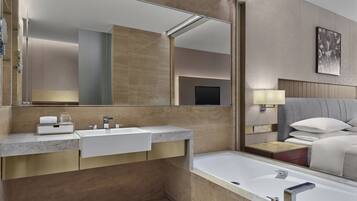 A modern bathroom with a large mirror, two sinks, a bathtub, and a bedside table with a lamp.